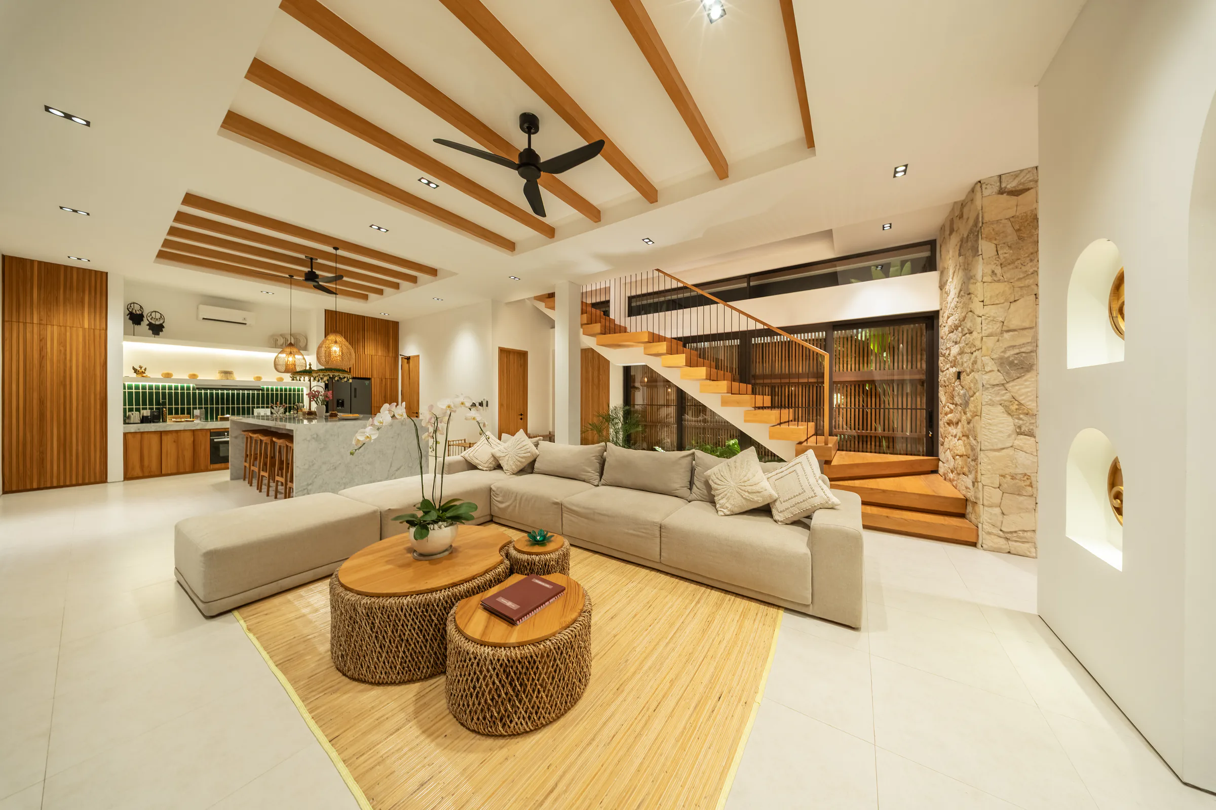 Living room with a long sofa under timber beams, beside a floating timber stair and a river stone wall
