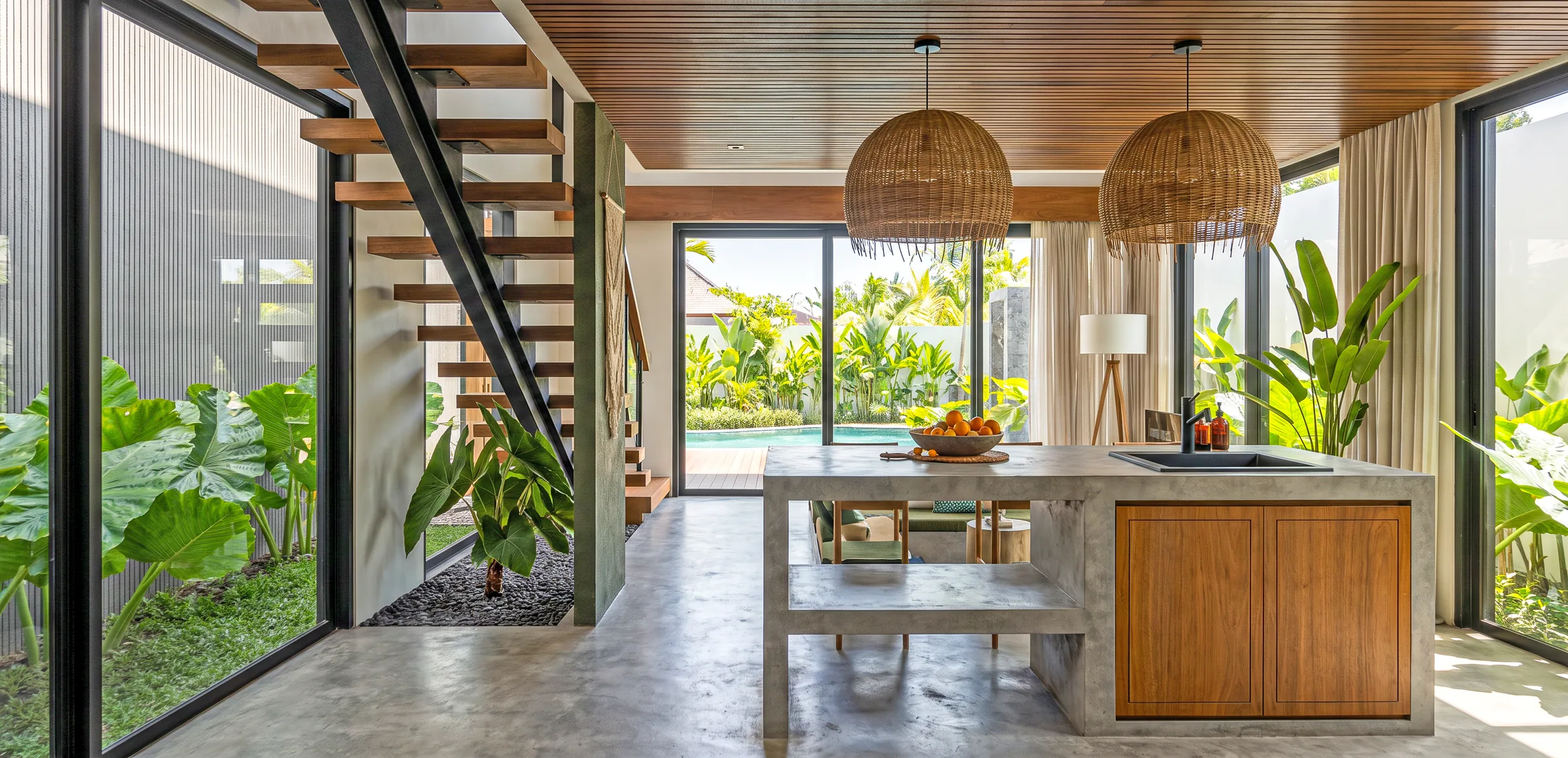 Kitchen with a concrete island and timber cabinets under woven pendants, between a steel stair and glass walls to the garden