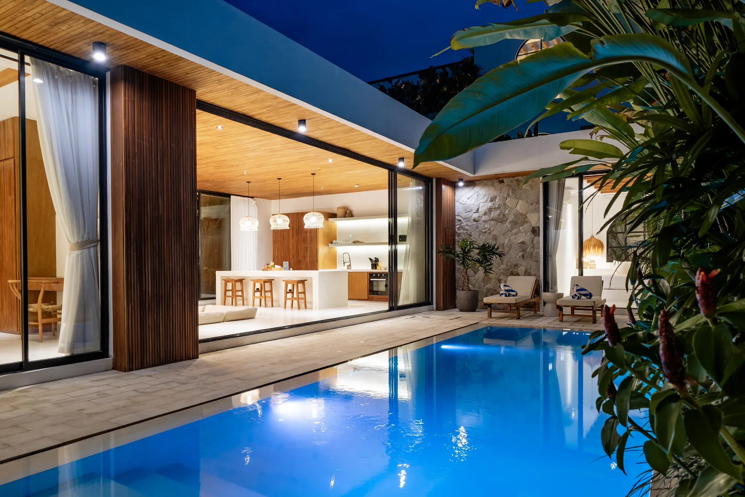 Casa Petak, a finished Balitecture villa in Canggu, at dusk: private pool, sun loungers set with fresh towels and the open-plan kitchen lit behind glass